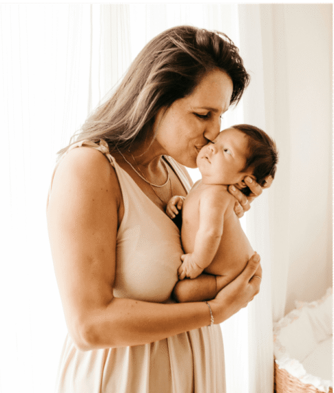 Mamá de pie cargando a su bebé y dándole un beso, representando la calma, seguridad y conexión que puede sentir al tener claridad sobre la alimentación en lactancia.